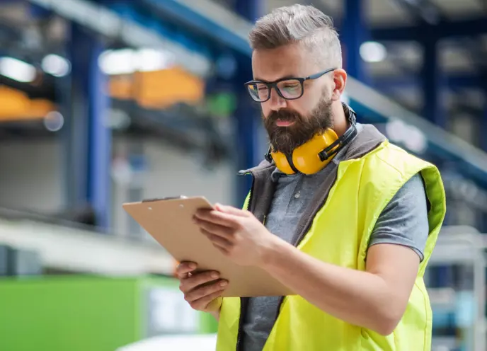 A man wearing a high-visibility vest and yellow ear protectors stands in an industrial setting, holding a clipboard and examining documents.